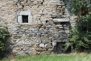 Casaforte Bozel a Villair (Morgex – Valle d’Aosta) lato est: Al di sotto del “marcapiano” e alla sinistra abbiamo l’inserimento a posteriori di una finestra quadrata dotata d’inferriata, la cui cornice è in pietra grigia e composta da quattro elementi; potrebbe o precorrere o imitare le “finestre a croce” cinquecentesche tipiche della Valle. Al centro abbiamo una piccola e rozza feritoia e alla destra vi è una caditoia “a cassetta” su beccatelli situata a meno di due metri da terra. 